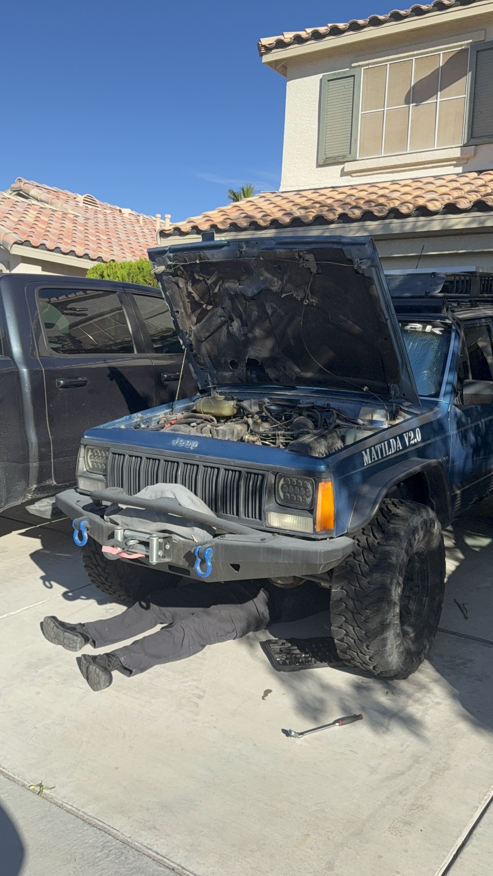 Mechanic working under a lifted blue Jeep Cherokee in a residential driveway